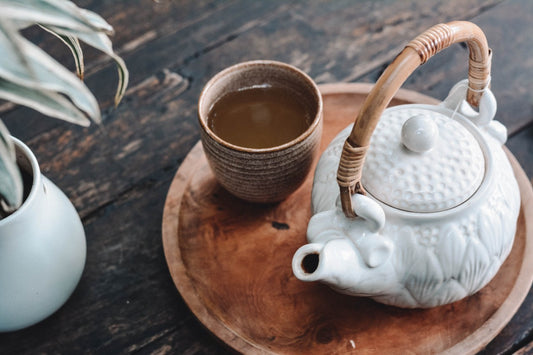 Tea cup full of liquid and a white teapot on a round timber serving tray